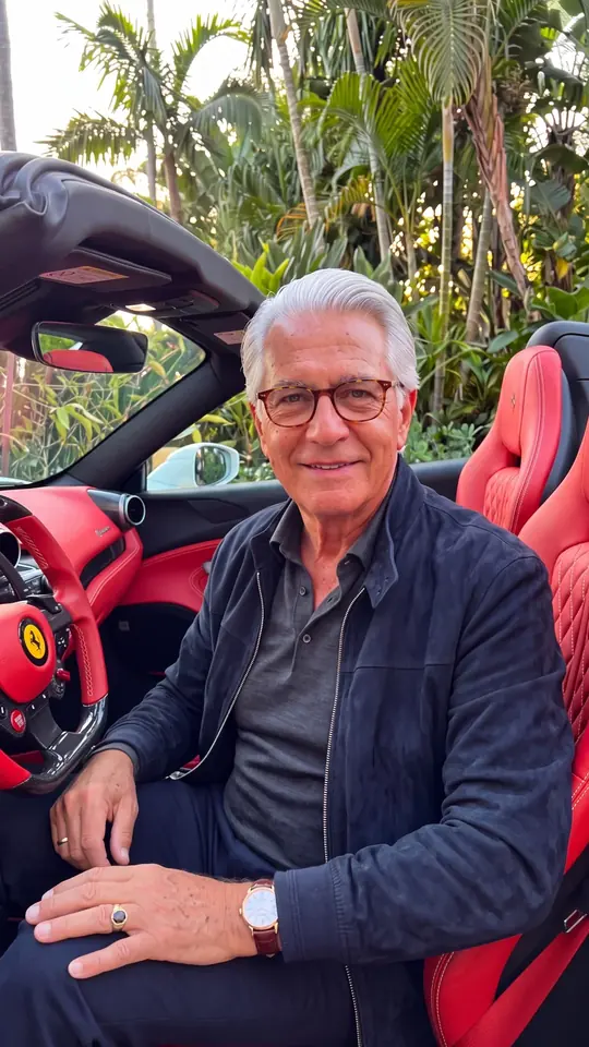 AI-generated character: a silver-haired man in a navy jacket seated in a red convertible among palm trees.